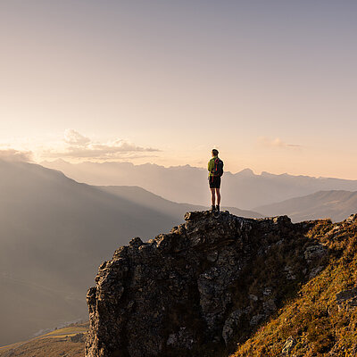 Marchkopf_5©Zillertaltourismus_PhillipGeisler Wanderin steht auf einem Felsen und blickt bei tief stehender Sonne über die alpine Berglandschaft.